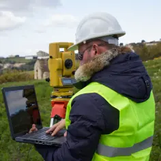 Geodeta w kamizelce odblaskowej i kasku pracuje z laptopem i tachimetrem. Dowiedz się, ile kosztuje wyznaczenie granicy działki.