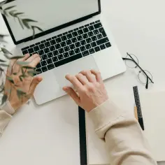 Ręce na klawiaturze laptopa, obok okulary i notatnik. Szybko sprawdzisz, jak sprawdzić czy księga wieczysta nie jest obciążona.