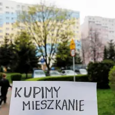 Znak "Kupimy mieszkanie" na tle bloków mieszkalnych. Co zrobić, gdy nie stać nas na mieszkanie? Szukamy rozwiązań.