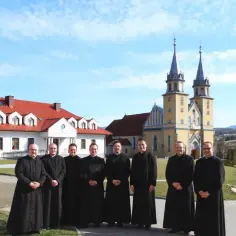 Grupa księży w czarnych szatach przed żółtym kościołem. Dyskutują, czy kościół płaci podatek od gruntu, podczas gdy słońce świeci.