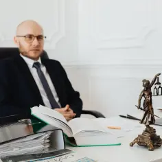 Prawnik w okularach siedzi przy biurku, obok stosu dokumentów i figury Temidy. Dowiedz się, ile kosztuje podział działki u notariusza.