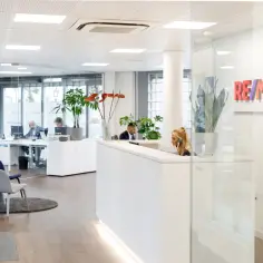 Nowoczesne biuro nieruchomości RE/MAX. Pracownicy obsługują klientów przy recepcji i biurkach.