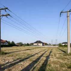 Dwa betonowe słupy z przewodami, linie wysokiego napięcia przecinają niebo nad polami i domami.