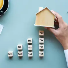Drewniany domek na kostkach z napisem "TAX", symbolizujący podatek od nieruchomości.