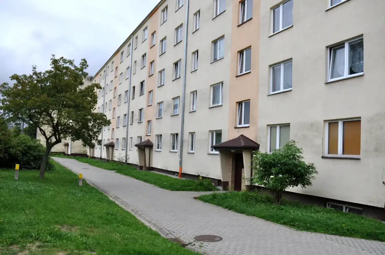 Długi blok mieszkalny z wieloma oknami i zielenią. Zastanawiasz się, ile trzeba zarabiać, żeby dostać mieszkanie komunalne?