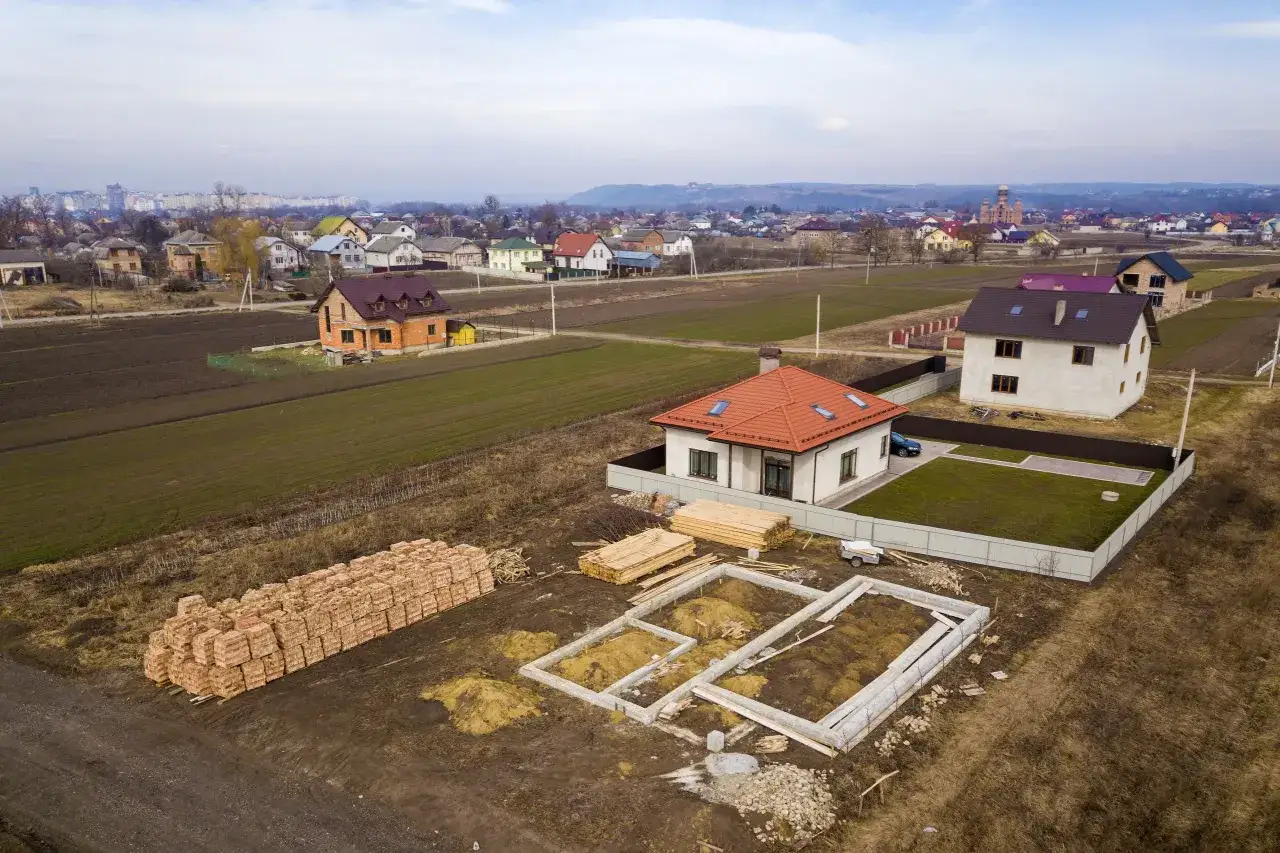 Widok z lotu ptaka na osiedle domów w budowie. Widać fundamenty, sterty cegieł i drewna, gotowe domy oraz pola uprawne. To idealne miejsce na jak duża działka pod budowę domu.