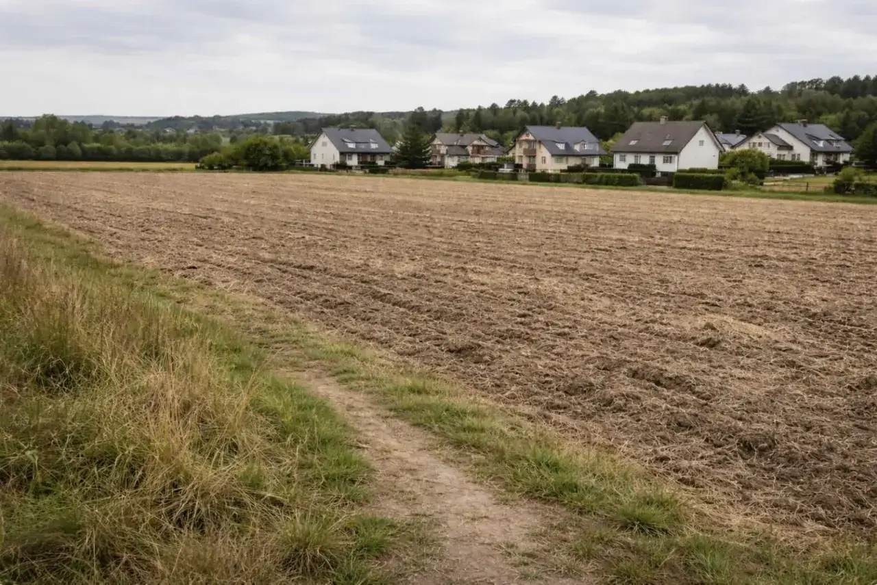 Pola uprawne i domy tworzą krajobraz działki rolno budowlanej. Ścieżka prowadzi przez zaoraną ziemię w kierunku zabudowań.