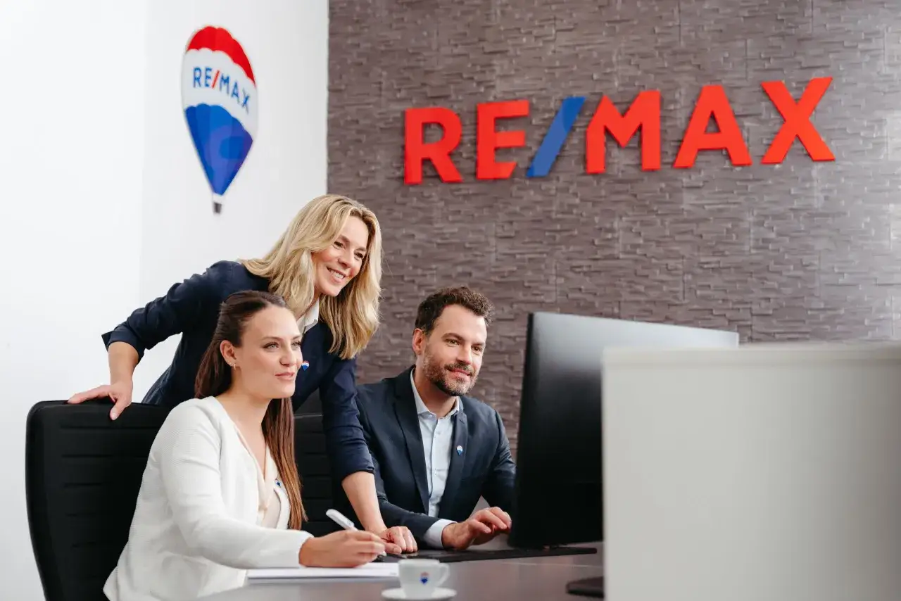 Zespół RE/MAX omawia strategię, jak założyć biuro nieruchomości. Na ścianie logo RE/MAX i balon.
