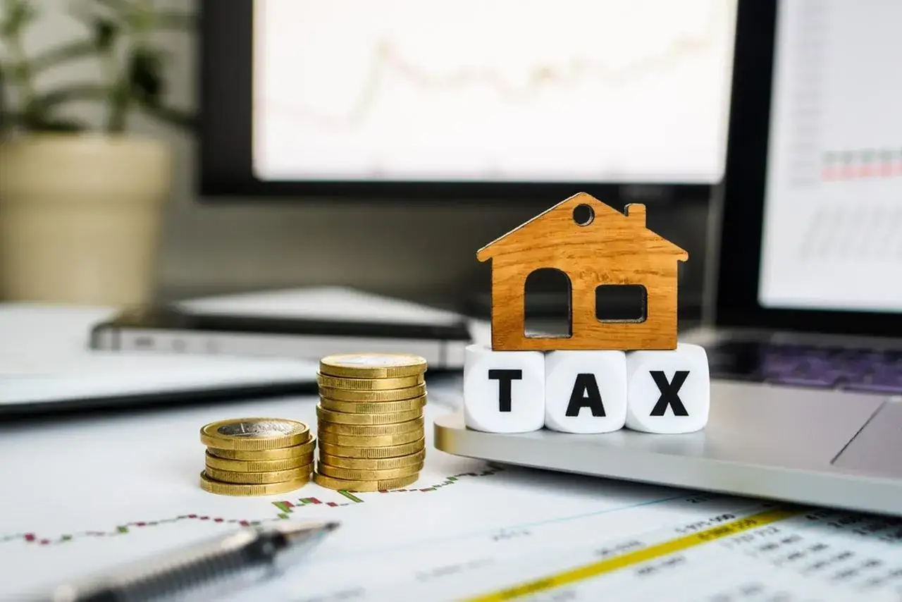 Drewniany domek na kostkach z napisem TAX, stosy monet i wykresy. Zastanawiasz się, ile wynosi podatek od wzbogacenia?