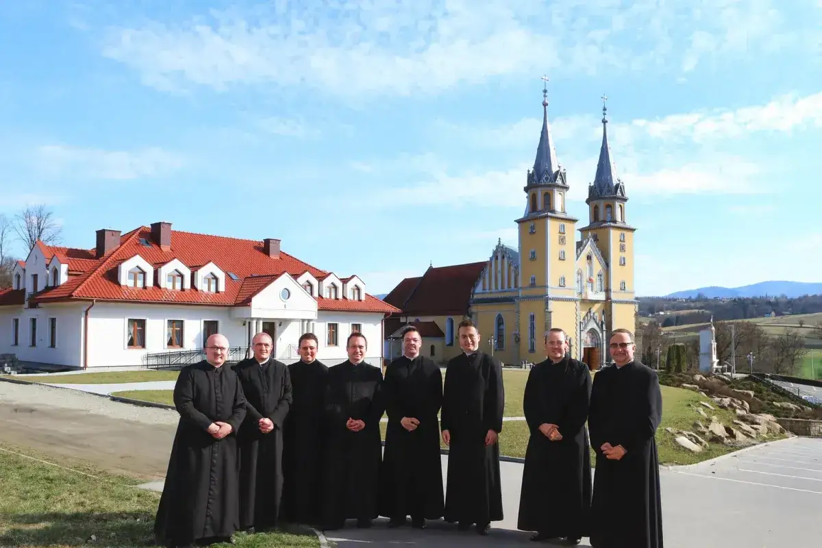 Grupa księży w czarnych szatach przed żółtym kościołem. Dyskutują, czy kościół płaci podatek od gruntu.