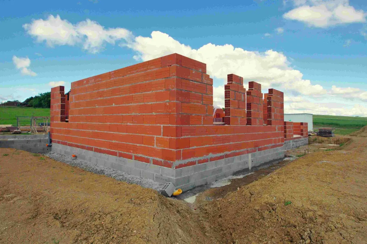 Wczesne etapy budowy domu: wznoszone ściany z cegły na fundamencie z bloczków betonowych, w tle zielone pole i błękitne niebo z chmurami.