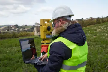 Geodeta w kamizelce odblaskowej i kasku pracuje z laptopem i tachimetrem. Dowiedz się, ile kosztuje wyznaczenie granicy działki.
