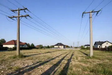 Dwa betonowe słupy z przewodami, linie wysokiego napięcia przecinają niebo nad polami i domami.