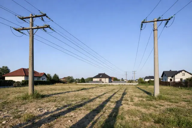 Dwa betonowe słupy z przewodami, linie wysokiego napięcia przecinają niebo nad polami i domami.